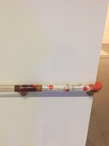 A plexiglass tube printed with orange patterns and gold text is mounted on a white wall at knee level, and curves around its corner. 
